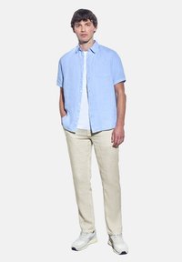 Chemise en lin légère à manches courtes de couleur bleu clair avec une poche poitrine, associée à un pantalon en lin blanc cassé et des baskets grises. Fond uni.