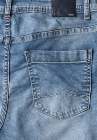 Tela de denim azul claro con una textura deslavada distintiva. Presenta un bolsillo trasero con costuras angulares y una etiqueta de la marca adjunta.