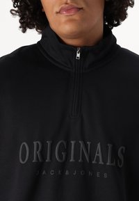 Crni dukserica s četvrtim zatvaračem s reljefnim natpisima "ORIGINALS" i "JACK&JONES" u mat finišu. Glatka tkanina i dizajn visokog ovratnika.