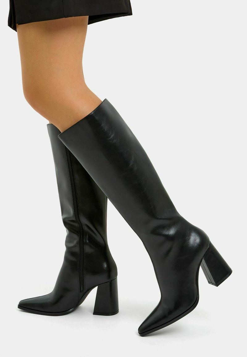 Femme portant des bottes en cuir noires montantes jusqu'au genou, à bouts pointus et talons blocs, associées à une jupe noire sur un fond uni.