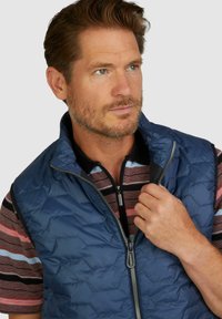 Marineblaue gesteppte Weste mit hohem Kragen, Frontreißverschluss und hexagonalem Muster. Getragen über einem gestreiften Poloshirt in Rot, Blau und Schwarz.