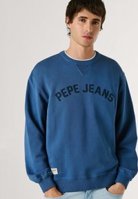 Mladý muž v modré mikině Pepe Jeans s rukama v kapsách světle modrých džínů, stojící před jednoduchým světlým pozadím.