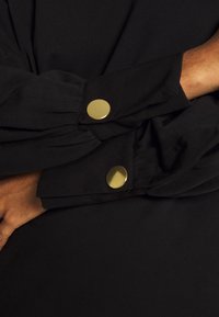 Indumento nero a maniche lunghe con polsini arrotolati, caratterizzato da bottoni a pressione in tono oro. Tessuto liscio con dettagli di cucitura sottili.