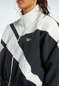 Svart och vit Reebok-jacka med dragkedja framtill, hög krage, elastisk midja och diagonalt färgblockdesign. Texturerad tyg.