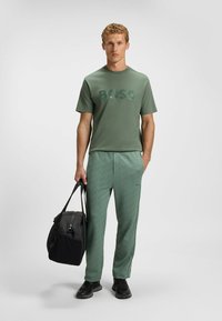 Grøn t-shirt med korte ærmer og tekstureret "BOSS" logo, parret med matchende mønstrede sweatpants. Modellen holder en sort duffel taske.