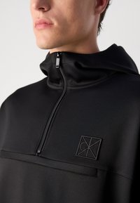 Čierna mikina na zips so zvýšeným golierom, textúrovanou tkaninou a malým vyšitým logom na hrudi. Hladký povrch a aerodynamický dizajn.