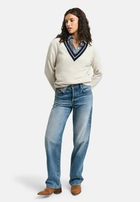 Cremfarbener V-Ausschnitt-Pullover mit marineblauen Akzenten, über einem blau-weiß gestreiften Hemd getragen, kombiniert mit hellblauen Jeans und braunen Stiefeletten.