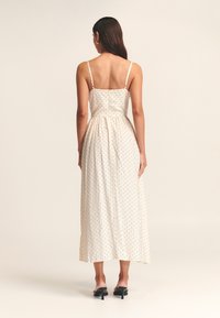 Light beige maxi dress with black polka dots, thin adjustable straps, fitted bodice, and gathered waist. Smooth fabric, ankle-length.