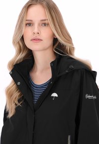 Zwarte waterdichte jas met ritssluiting, knoopaccenten en een wit parapluelogo op de borst. Gedragen over een blauwgestreept overhemd.