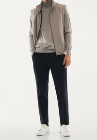 Herrenoutfit mit einem hellgrauen Zip-Up-Weste, grauem Rollkragenpullover, dunklen Hosen und weißen Sneakers. Glatter Stoff, figurbetontes Design und neutrale Farben.