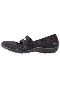 Zapato plano negro con tejido de punto texturizado y una correa de velcro. Cuenta con una suela de goma flexible con detalles de tracción.