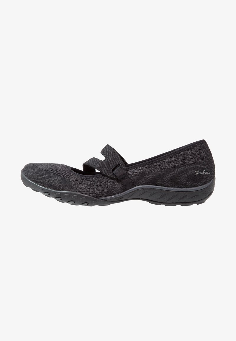 Zapato plano negro con tejido de punto texturizado y una correa de velcro. Cuenta con una suela de goma flexible con detalles de tracción.