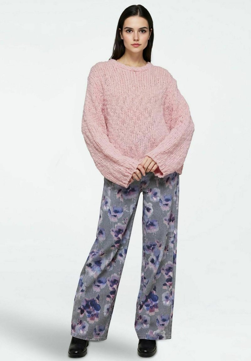 Maglione rosa oversize lavorato a maglia con maniche larghe, abbinato a pantaloni grigi a gamba larga con un motivo floreale viola e stivaletti neri alla caviglia.