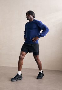 Felpa blu, pantaloni corti neri e sneaker neri. Il tessuto è morbido, con polsini a coste e cuciture ben visibili. Uno stile minimalista.