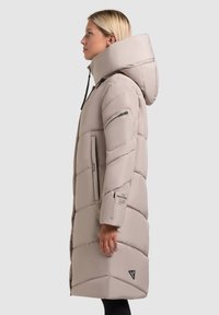 Vrouw in een lange beige gewatteerde winterjas met een hoge kraag, capuchon, ritszak op de arm en zijzakken, staand in profiel.