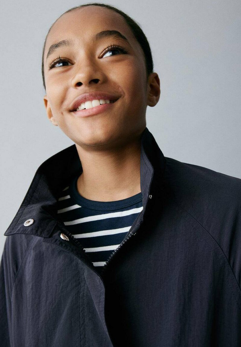 Chaqueta navy con cuello alto y botones a presión, llevada sobre una camisa de rayas en azul y blanco. Tejido suave, diseño casual, mínimas detalles.