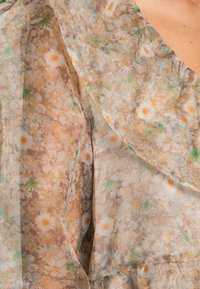 Blouse en tissu à imprimé floral beige clair avec des accents orange et vert, présentant un décolleté drapé et une texture douce et transparente.