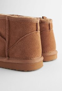 Bruna mockalboots med en mjuk pälsfoder, med stickade detaljer och en strukturerad gummisula, visar en närbild av baksidan.