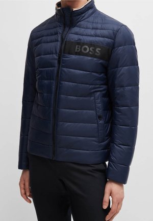 Mand iført en marineblå quiltet jakke med en sort horisontal stribe og teksten "BOSS" på tværs af brystet, lynet op, stående stille.