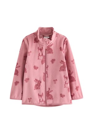 Glimlachend roodharig kind met een roze jas met vosjespatroon, blauwe jeans, zachte oorwarmers en kleurrijke sneakers, staand tegen een effen achtergrond.