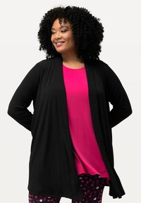 Schwarze langärmlige Strickjacke, offene Vorderseite, über einem leuchtend pinken Oberteil und gemusterten Leggings mit Kreisen in verschiedenen Schattierungen von Pink und Lila.