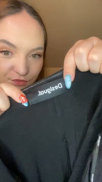 Prenda negra con la etiqueta "Desigual" mostrada de manera prominente, con cierre y textura suave. Primer plano con mano visible sosteniendo la tela.