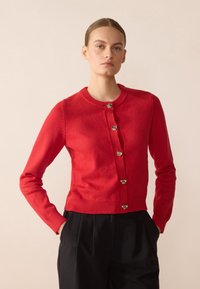 Cardigan en tricot rouge avec encolure ronde, manches longues et boutons en ton or. Ourlet et poignets côtelés texturés, offrant une silhouette ajustée.