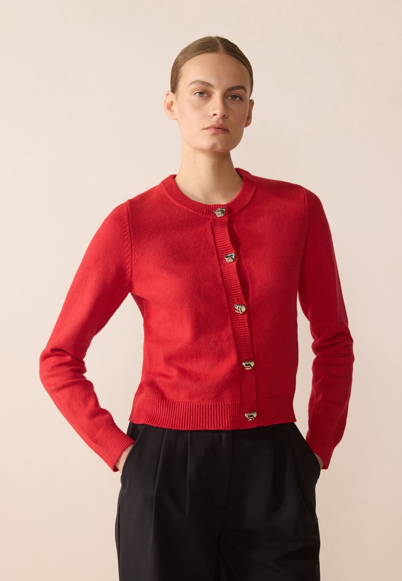 Cardigan en tricot rouge avec encolure ronde, manches longues et boutons en ton or. Ourlet et poignets côtelés texturés, offrant une silhouette ajustée.