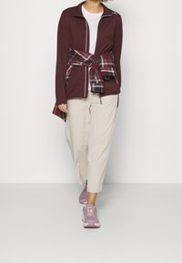 Veste zippée bordeaux avec des liserés blancs, associée à un t-shirt blanc, un pantalon beige et des baskets violet clair. Chemise à carreaux nouée autour de la taille.