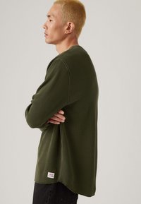 Groene trui met wafelstructuur en lange mouwen, ruime pasvorm, met een afgeronde onderkant en een klein merkje aan de onderzijde links.