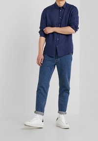 Uomo in camicia blu navy arrotolata, jeans blu risvoltati alle caviglie, calzini bianchi e sneakers bianche in piedi contro uno sfondo grigio semplice.