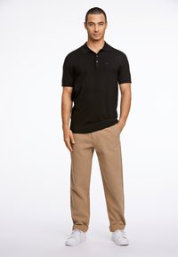 Polo negro de manga corta con cuello, con un logo sutil, combinado con pantalones chinos beige y zapatillas blancas. Diseño simple y ajustado.