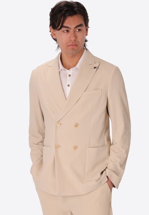 Uomo con capelli scuri che indossa una giacca doppiopetto beige e pantaloni abbinati con le mani in tasca, che guarda di lato.