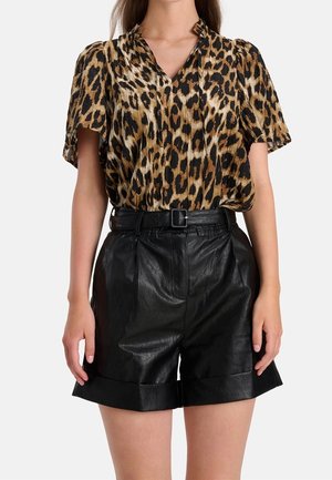 Vrouw draagt een korte mouwen blouse met luipaardprint, ingestopt in een hooggetailleerde zwarte leren short met een zwarte riem.