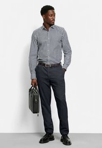 Chemise rayée à boutons bleu marine et blanche, associée à un pantalon foncé. Tenant une mallette grise, portant des chaussures noires, debout devant un fond uni.