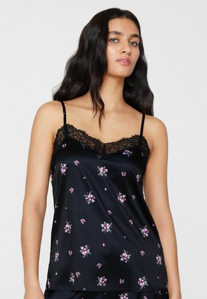 Młoda kobieta z długimi ciemnymi włosami, ubrana w czarną satynową koszulkę na ramiączkach z małymi różowymi kwiatowymi nadrukami i czarną koronkową lamówką.