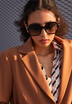 Blazer marrón con solapas anchas sobre una camisa estampada con diseños geométricos en negro, blanco y rojo. Unas grandes gafas de sol negras completan el look.