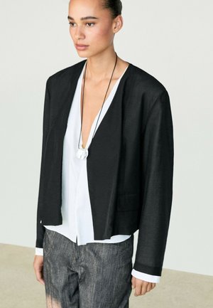 Femme aux cheveux attachés portant une veste noire ouverte sur un chemisier blanc à col en V profond et un pantalon gris, avec un collier pendentif rond.
