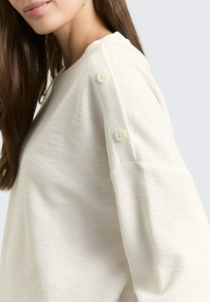 Vrouw draagt een witte, lange mouwen gebreide top met twee witte knopen langs de schoudernaad, zichtbaar lang bruin haar.