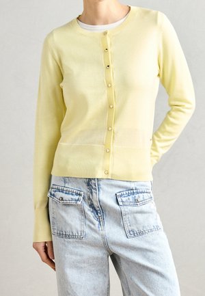 Personne portant un cardigan à boutons jaune clair et un jean denim bleu clair avec poches à rabat à l'avant. Une main repose dans le dos.