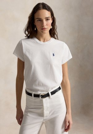 Kobieta w białej koszulce z małym niebieskim logo na piersi, białych spodniach, czarnym pasku i kolczykach w kształcie muszli, stojąca na neutralnym tle.