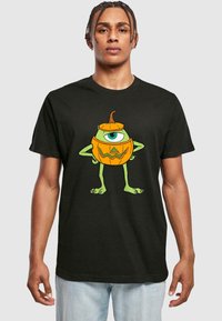 Black cotton T-shirt featuring a green character with a pumpkin design. The character has one eye, a jagged mouth, and stands with arms akimbo.