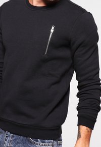 Sudadera negra con mangas largas, escote redondo y un pequeño bolsillo con cremallera plateada en el pecho. Tejido suave con textura lisa.