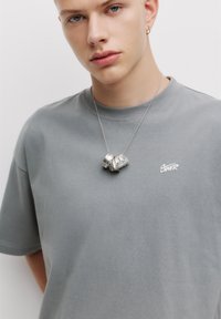 Grå bomulds t-shirt med korte ærmer, med et lille hvidt logo. En sølvhalskæde med snoede og strukturerede designs ligger på brystet.