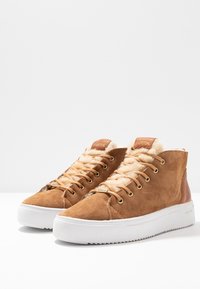 Brown suede high-top sneakers with a soft tan lining, gold eyelets, white rubber soles, and a textured pattern on the sole.
