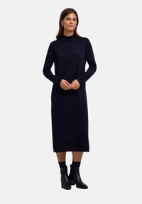Navyblaues Langarmkleid mit einem hohen Kragen, taillierter Silhouette, weichem Material, das bis unter die Knie reicht, kombiniert mit schwarzen Stiefeletten.