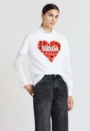 T-shirt bianco con una grafica di un cuore floreale rosso e il testo "NIRVANA", abbinato a jeans grigi scuri a vita alta. Maniche lunghe visibili sotto.