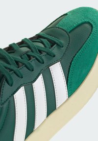 Grüner Sneaker mit weißen Streifen, Wildleder- und Ledertexturen, grünen Schnürsenkeln und beige-farbener Sohle, Nahaufnahme von Vorder- und Seite.