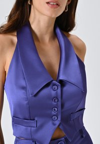 Top halter in satin viola con scollatura a V profonda, ampio colletto e chiusura con sei bottoni. Presenta tasche laterali e una texture liscia.