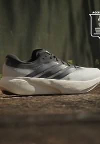 Chaussure de course Adidas gris clair et noir avec une semelle blanche épaisse étiquetée « DENSITYSTRIKE » sur un sol en terre en extérieur.